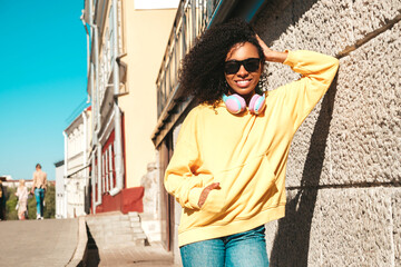 Beautiful black woman with afro curls hairstyle.Smiling model in yellow hoodie.Sexy carefree female enjoying listening music in wireless headphones.Posing on street background near wall in sunglasses