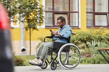 Fototapeta na wymiar Content young African-American man with ill legs sitting in wheelchair and reading sms on phone outdoors