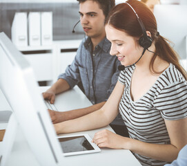 Casual dressed young woman using headset and computer while talking with customers online. Group of operators at work in sunny office. Call center