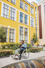 Positive young African-American man with paralyzed legs sitting in wheelchair and recording audio message on phone against sanatorium building