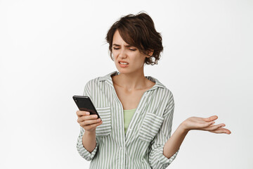 Fototapeta premium Frustrated young woman looking at mobile phone screen complicated, shrugging and frowning disappointed, standing over white background