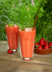 Two glasses with strawberry smoothie with mint leaves and glass straws on a wooden table in the garden