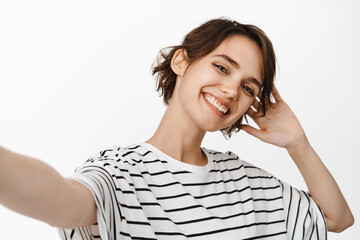 Fototapeta premium Beautiful brunette woman taking selfie, extend hand, holding camera and posing, standing in t-shirt against white background
