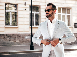 Portrait of handsome confident stylish hipster lambersexual model.Modern man dressed in elegant white suit. Fashion male posing in the street background in Europe city at sunset. In sunglasses