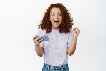 Excited girl winner, redhead woman holding mobile phone and looking amazed as if winning, become champion in video game, white background