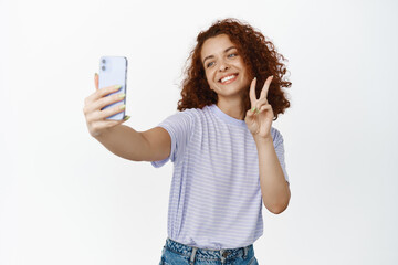 Happy beautiful girl taking selfie on smartphone with peace sign, smiling at mobile phone camera, white background