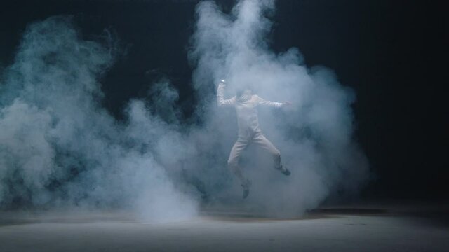 Professional fencer in protective uniform practicing maneuvers with sword alone in dark studio . Fencing demonstration on black smoked background . Shot on Arri Alexa cinema camera in slow motion .	