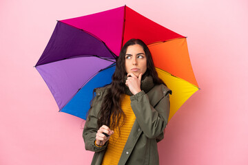 Young woman holding an umbrella isolated on pink background having doubts