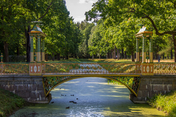 Fototapeta na wymiar an elegant bridge over the canal in the Tsarskoye Selo Park and bright green trees and grass on a sunny summer day. Concept-walking and tourism in Leningrad region Russia