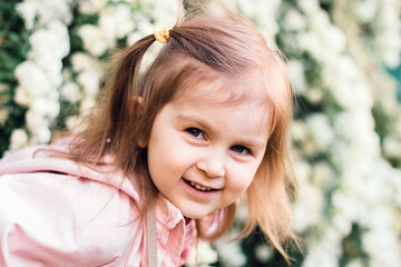 Fototapeta premium Cute smiling little caucasian girl walking in park outdoors and looking at camera. Nature background with blooming bush.