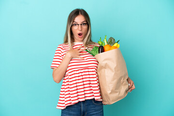 Young woman holding a grocery shopping bag isolated on blue background surprised and shocked while looking right