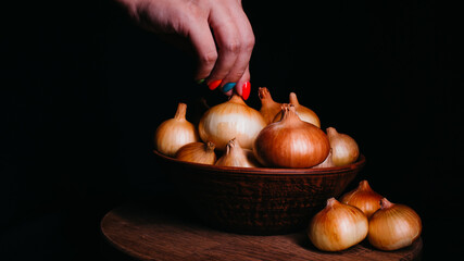 Pile of whole bulbs of raw onion in ceramic bowl on table. Woman's hand takes bulb of onion from bowl.