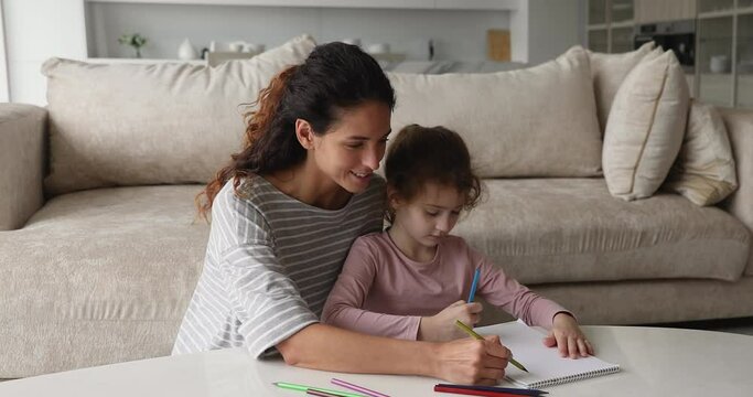 Caring affectionate young mother teaching small cute kid daughter drawing pictures in paper album with colored pencils, developing creativity, improving art skills, enjoying domestic hobby activity.