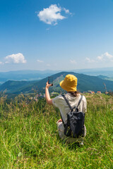 Hipster traveler in a hat and backpack, resting in nature and using the Internet on a smartphone. A tourist girl photographs a panorama of the mountains by a mobile phone. Mountain beautiful landscape