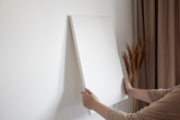 Blank canvas in female hands, picture mockup. Woman hanging canvas on white wall