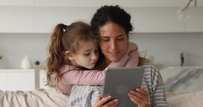 Loving small preteen kid girl embracing beautiful young mommy or nanny, using digital computer tablet together at home, web surfing information, playing games or shopping in internet store together.