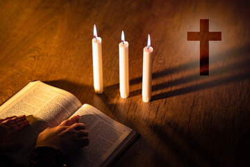 Priest hands on bible. Burning candles next to Christian cross. Cross symbolizes Catholicism or...