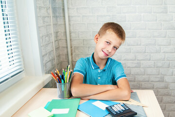 Smiling boy is sitting at school desk. Stationery is laid out on table near the child.