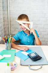Smiling boy is playing with ruler sitting at school desk. Stationery is laid out on table near child.