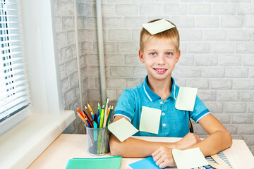 Laughing boy is playing with stickers while sitting at school desk. Stationery is laid out on table near the child. Holidays. Back to school.