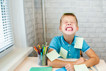 Laughing boy is playing with stickers while sitting at school desk. Stationery is laid out on table near the child. Holidays. Back to school.