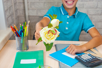 The hand of a boy who holds out a rose to the teacher. A child sitting at a school desk. Stationery is laid out on the table near the child. Holidays. Back to school.