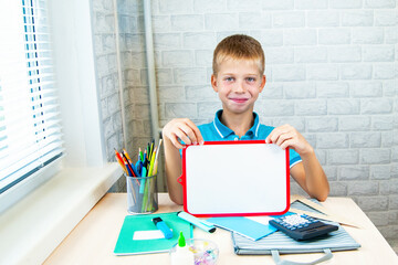 A boy student is sitting at a desk and holding a marker board in his hands. Office supplies are laid out on the table. Back to school.