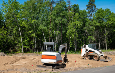 A mini excavator with hydraulic scoop sits on the dirt at a new home construction lot, with trees in back of site.