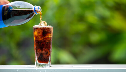 Cola pouring in to glass over sunlight and natural green background.Cola in glass with ice.