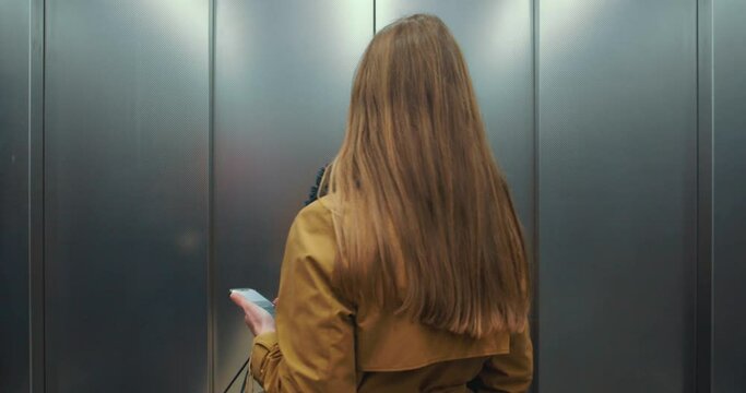 Young beautiful business woman in face mask with smartphone, shopping bags riding elevator, leaving into yellow hall.