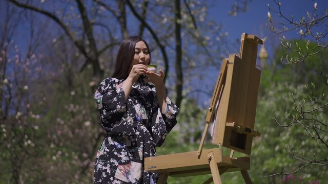 Gorgeous slim Asian woman in kimono drinking tea standing at easel in sunny spring park outdoors. Portrait of talented inspired female painter enjoying hobby in sakura garden outdoors. Beauty concept