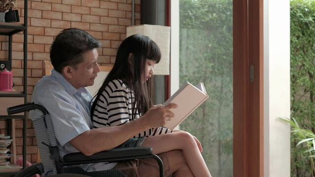 Granddaughter sat on the old Asian grandfather's lap, disabled and retired in a wheelchair in the house, while happily reading book together during the vacation. Love for a relationship lifestyle.