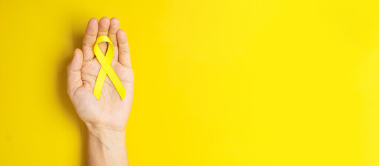 hand holding Yellow Ribbon on yellow background for supporting people living and illness. September Suicide prevention day, Childhood Cancer Awareness month and World cancer day concept - obrazy, fototapety, plakaty
