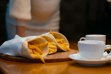 french fries in pita bread in a paper wrapper, packaging lies on the dining table in a dark room in the evening against the background of the belly of a pregnant girl, a woman and tea or coffee mugs