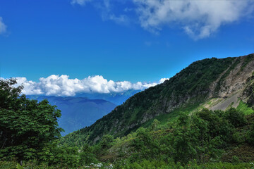 The mountain slopes are covered with green vegetation. Picturesque cumulus clouds can be seen above the peaks of the ridge. Blue sky. A sunny summer day. Caucasus. Krasnaya Polyana