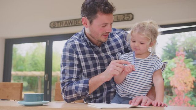 Father at home at kitchen counter helping young daughter to draw picture in book - shot in slow motion
