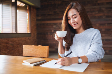 A beautiful young asian woman drinking coffee and writing on a blank notebook on the table