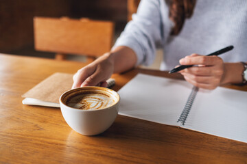 Fototapeta na wymiar Closeup image of a woman drinking coffee and writing on a blank notebook on the table