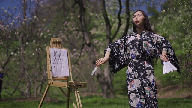 Confident gorgeous Japanese woman in kimono dancing with fans at painting easel in blooming spring park. Portrait of artistic Asian lady enjoying sunny day outdoors. Slow motion