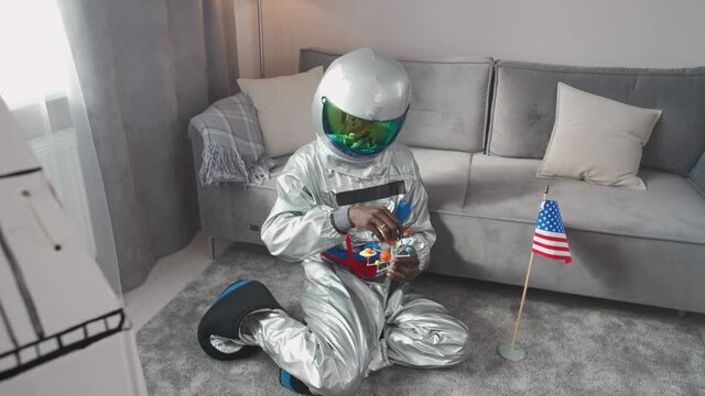 Afro american man in an astronaut costume sitting on the floor in living room at home and playing with a toy model of the solar system, man on self-isolation, the flag of USA in the foreground, 4k