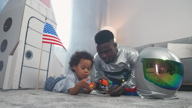 Afro american father and son play in the living room at home, man in a suit of an american astronaut lying on the floor with her son, boy playing with a toy model of the solar system, study of