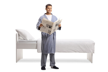 Young man with a newspaper wearing a bathrobe and standing next to a bed