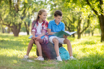 Fototapeta premium Cheerful boy and girl sitting on stump in park