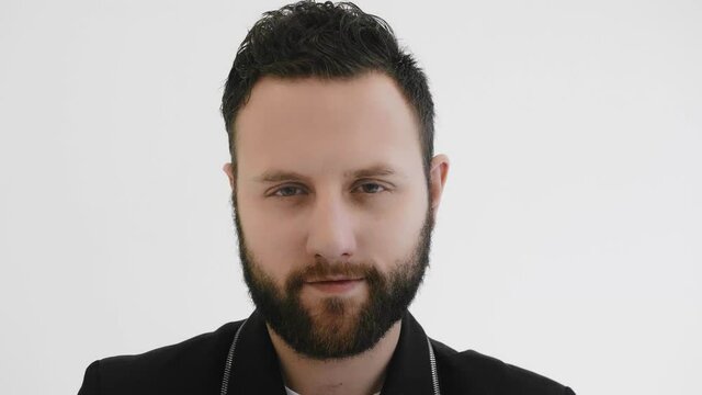 Fashionable stylish man moves his eyes around. Portrait of bearded brutal guy looking to camera. Trendy fancy person narrowed his eyes. Black jacket. Chic, modish. White background in studio. Closeup