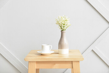 Vase with lily-of-the-valley flowers and cup of coffee on table near light wall