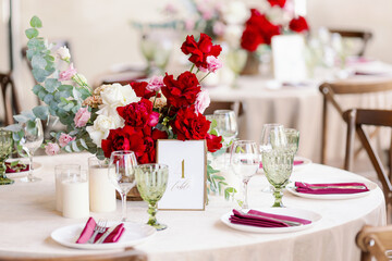 Decoration of the wedding table with red and pink flowers on a white tablecloth. Green glass goblets and framed table number.