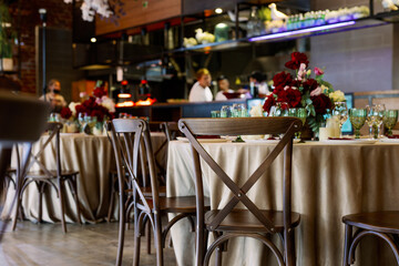 Dinner party indoors with served tables in a restaurant with red flowers and wooden vintage chairs in rustic boho style