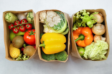 three paper bags with fresh vegetables and fruits on a light table, top view