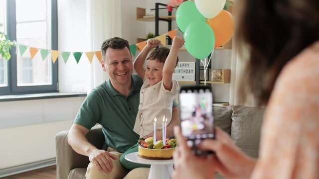 family, holidays and people concept - mother with smartphone photographing happy father and little son with birthday cake sitting on sofa at home party