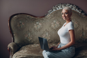 Millenial young woman with short blonde hair portrait sitting and smiling with laptop on vintage sofa © primipil